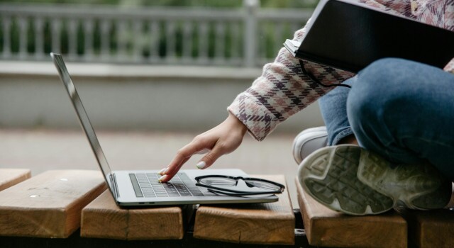PersoninBlueDenimJeansandPlaidShirtSittingonWoodenBench singLaptop