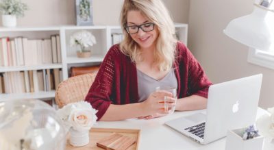 WomansmilingholdingglassmugsittingbesidetablewithMacBook